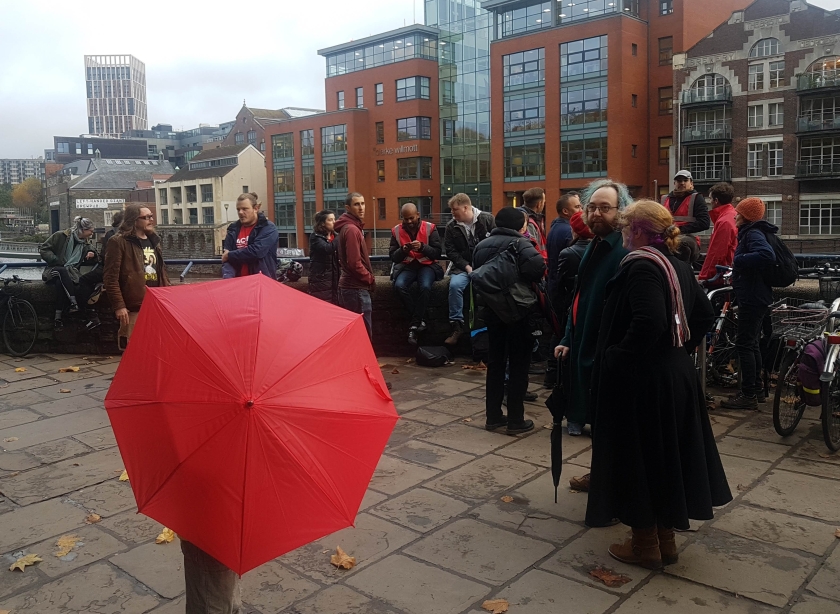 A group of around 20 people can be seen. Several are wearing red tabards and one has a red umbrella.