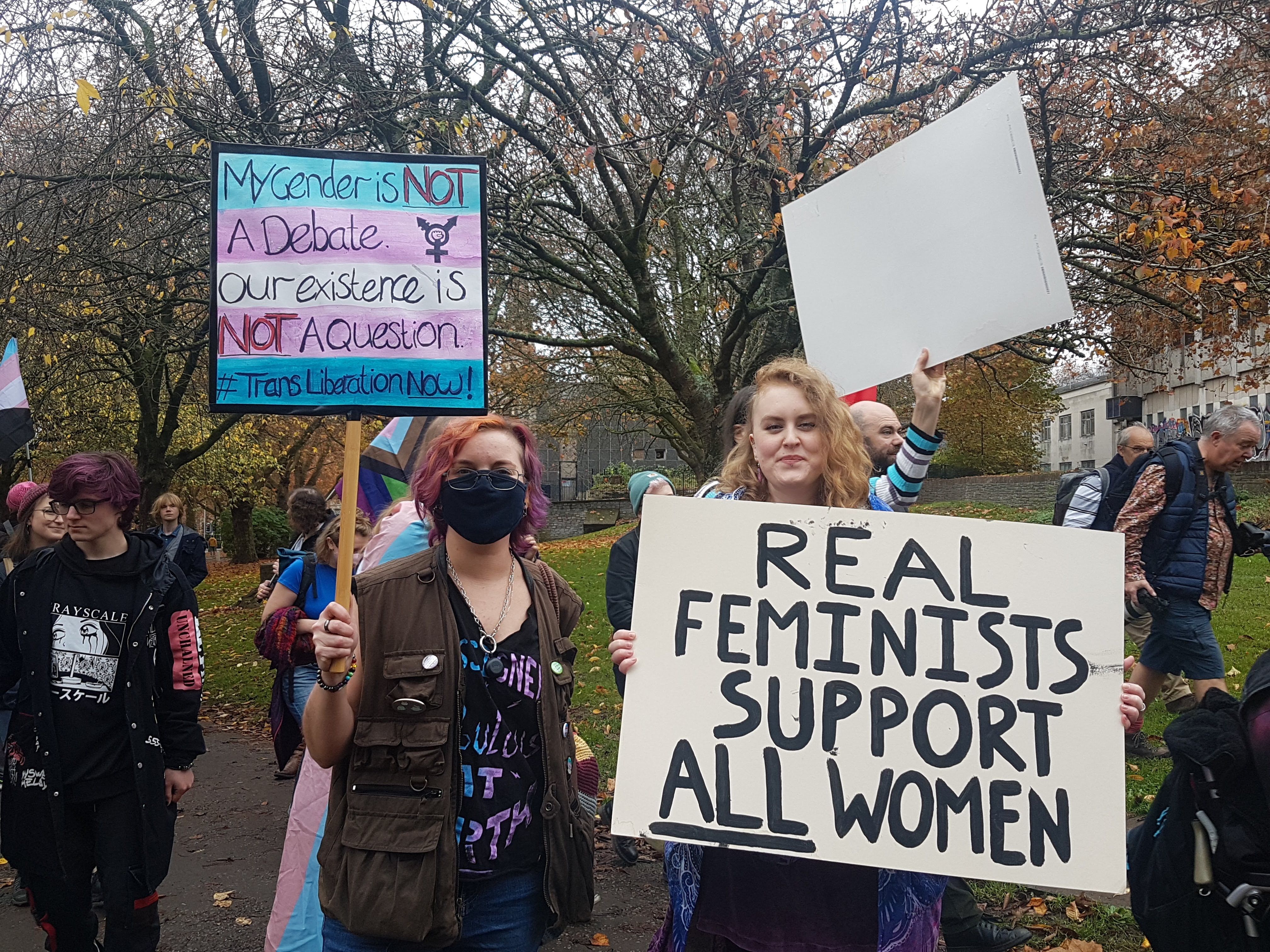 Two protesters hold placards saying "Real feminists support all women." and "My gender is not a debate."