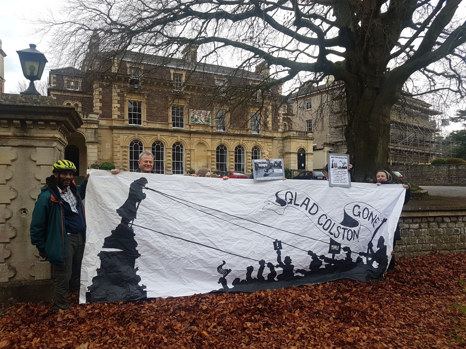 Seven people hold a large white banner with an image of Colston's statue being toppled. In theSeven people hold a large white banner with an image of Colston's statue being toppled. In the background is a mansion house. background is a mansion house.