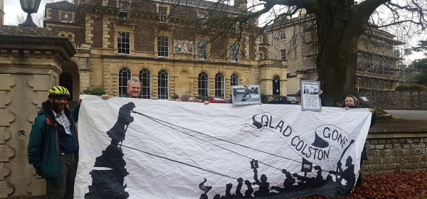 Seven people hold a large white banner with an image of Colston's statue being toppled. In theSeven people hold a large white banner with an image of Colston's statue being toppled. In the background is a mansion house. background is a mansion house.