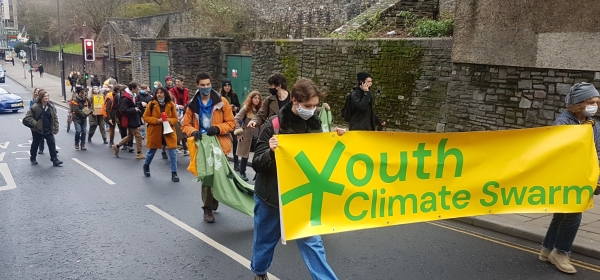 Protesters march in the street. At their head is a banner saying "Youth Climate Swarm"
