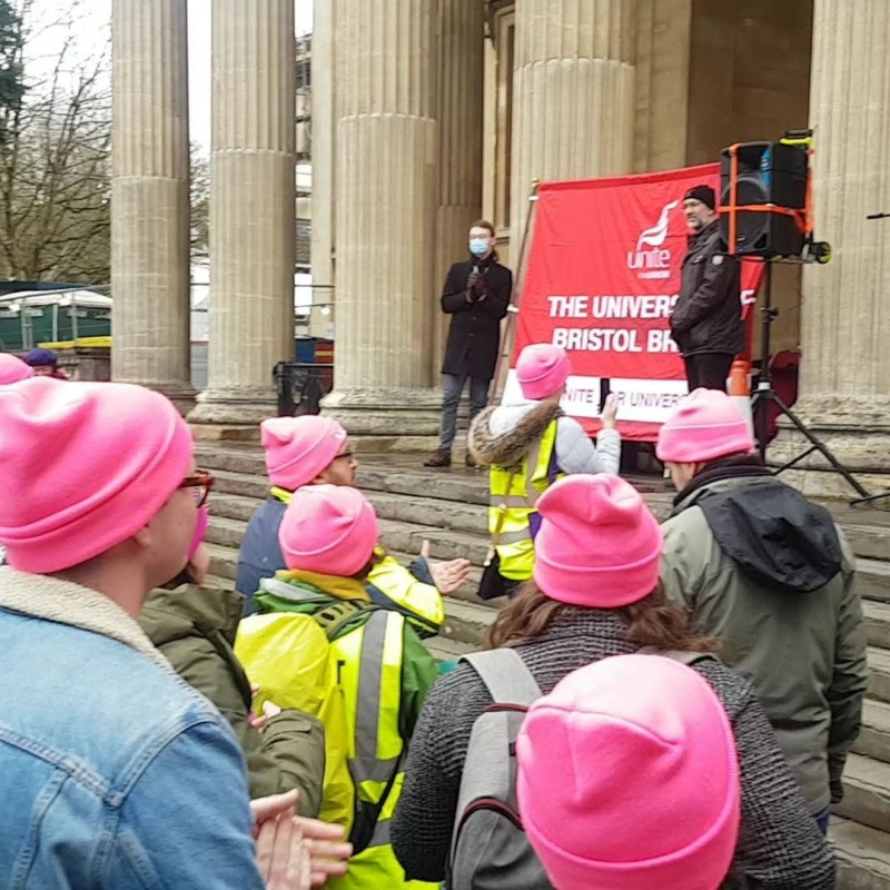 A crowd of people listen to a speaker. Many are wearing bright pink hats.