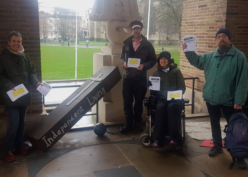 Four people pose holding leaflets. A mock coffin says "Independent Living RIP"