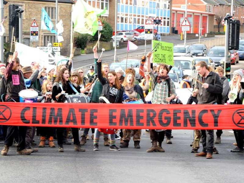 A group of around 12 people drum behind a banner reading "Climate Emergency"
