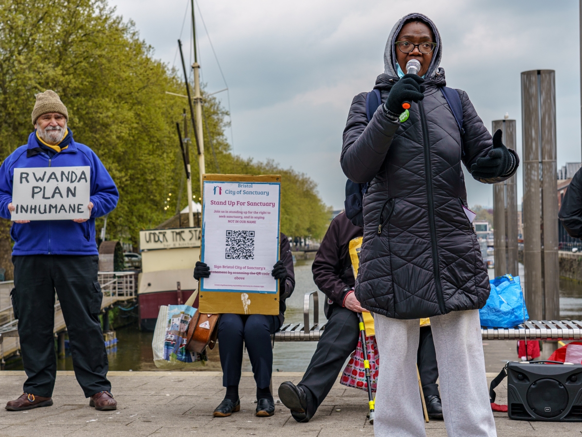 A woman holds a microphone whilst behind her a man holds a placard reading "Rwanda Plan Inhumane"